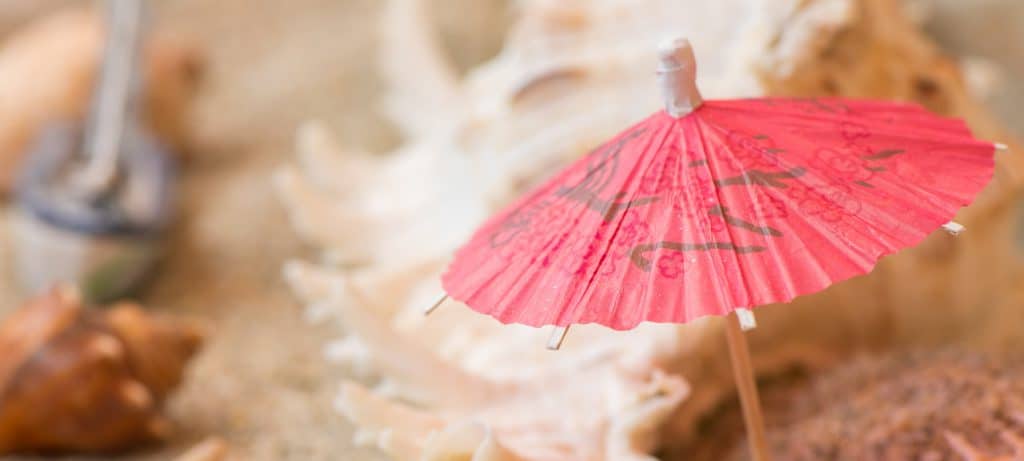 Ein rosa Cocktailschirmchen aus Papier steckt im Sand, umgeben von Muscheln und verschwommenen Strandobjekten im Hintergrund.