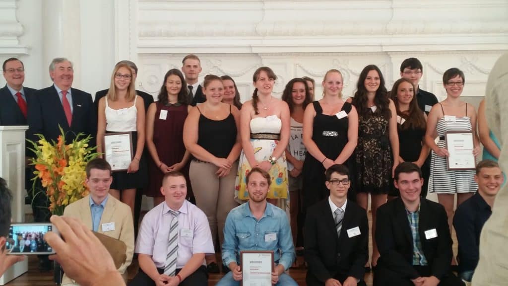 Eine Gruppe junger Erwachsener und einige ältere Menschen posieren im Innenbereich für ein formelles Foto. Einige halten Zertifikate in den Händen, die meisten tragen Namensschilder. Links sind Blumen arrangiert, und im Vordergrund macht eine Person ein Foto.
