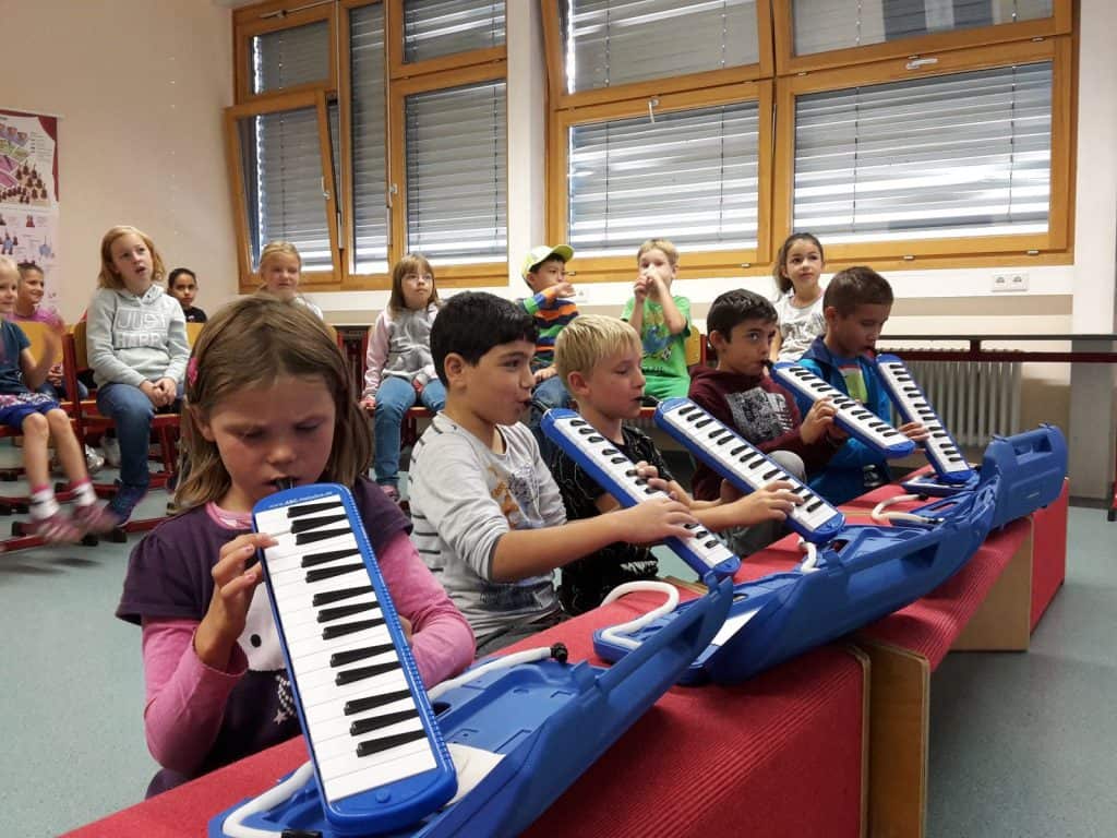 Eine Gruppe von Kindern sitzt in einem Klassenzimmer und spielt blaue Melodicas, während andere Kinder im Hintergrund zuschauen. Der Raum hat große Fenster mit Jalousien, und die Kinder scheinen sich auf ihre Instrumente zu konzentrieren.