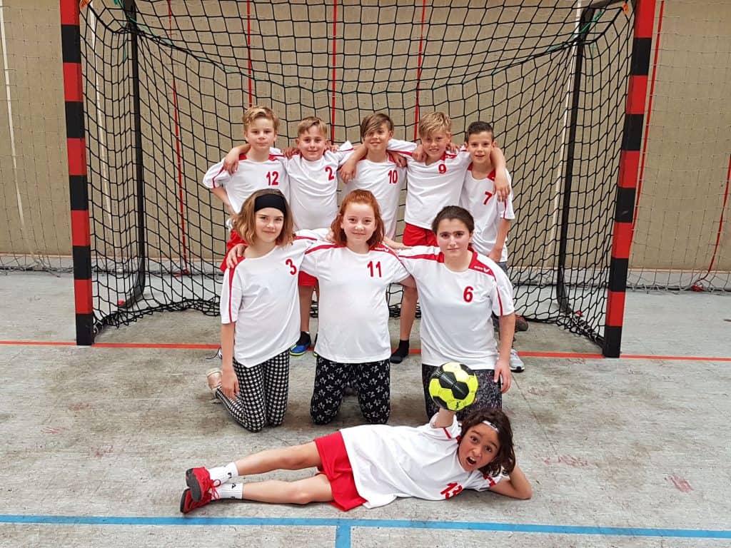 Eine Jugendhandballmannschaft aus neun Kindern in weiß-roten Trikots posiert vor einem Tornetz. Acht Kinder stehen und knien, ein Kind liegt mit einem Ball in der Hand davor. Alle lächeln in die Kamera.