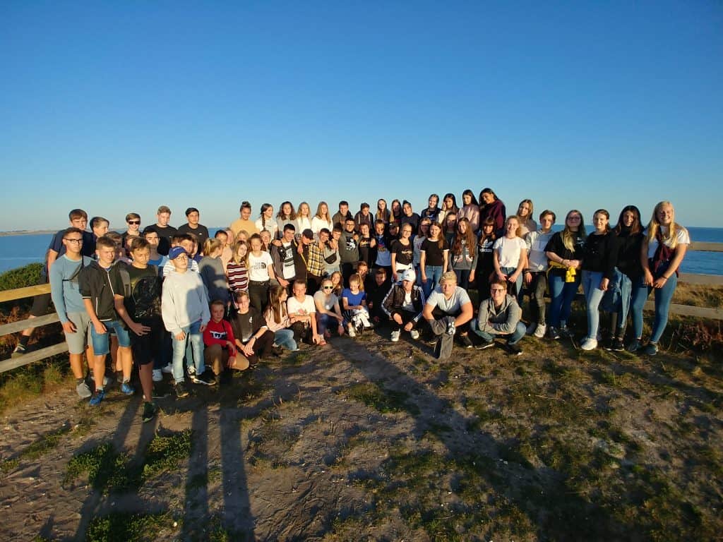 Eine große Gruppe von Teenagern und jungen Erwachsenen posiert an einem sonnigen Tag gemeinsam im Freien. Sie stehen auf Gras in der Nähe eines Holzzauns mit dem Meer und dem blauen Himmel im Hintergrund.