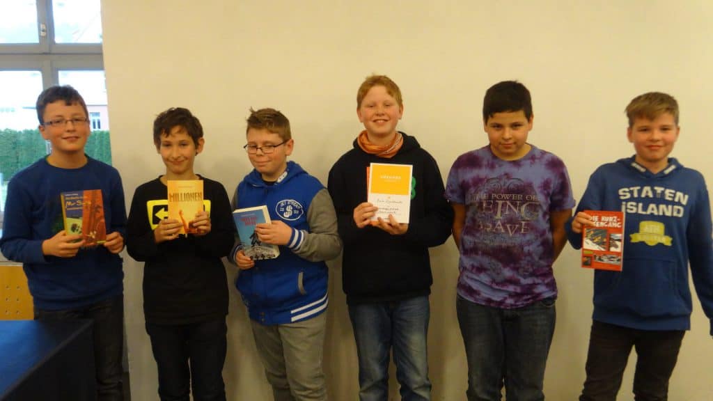 Sechs Jungen stehen nebeneinander vor einer weißen Wand, jeder hält ein Buch in der Hand und lächelt in die Kamera. Sie scheinen an einer Schul- oder Bibliotheksveranstaltung teilzunehmen.