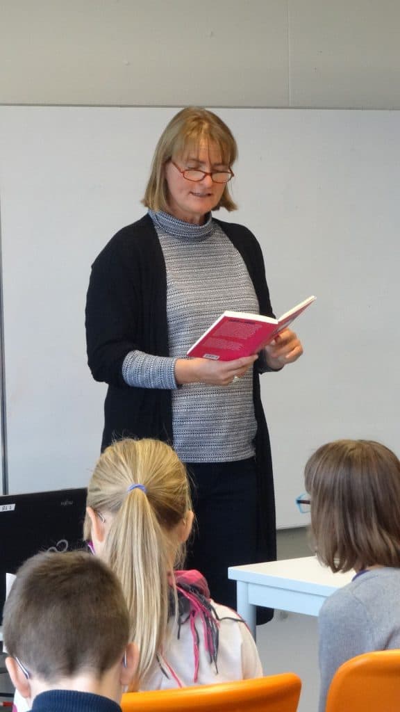 Eine Frau steht lächelnd vor einem Klassenzimmer, hält ein offenes Buch in der Hand, während mehrere Kinder ihr gegenüber sitzen und zuhören. Hinter ihr befindet sich eine Tafel, und im Vordergrund sitzen Schüler an Tischen.