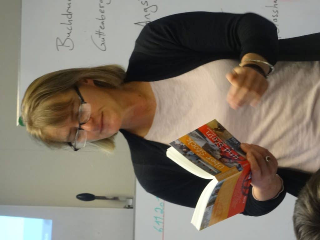Eine Frau mit Brille liest vor einer mit handschriftlichen Notizen bedeckten Tafel aus einem Buch. Sie trägt eine schwarze Strickjacke über einem hellen Hemd und scheint einer Gruppe etwas zu erklären.