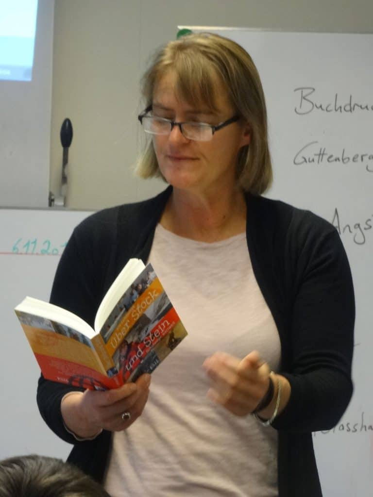 Eine Frau mit blonden Haaren und Brille liest in einem Klassenzimmer vor einer weißen Tafel mit deutscher Schrift. Sie trägt ein helles Hemd und eine dunkle Strickjacke.