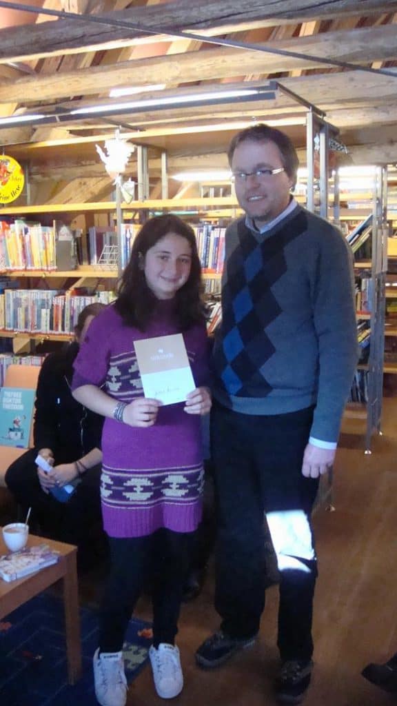 Ein junges Mädchen mit einem Zertifikat steht neben einem erwachsenen Mann in einer Bibliothek. Beide lächeln in die Kamera. Im Hintergrund sind Bücherregale voller Bücher und Holzbalken zu sehen.
