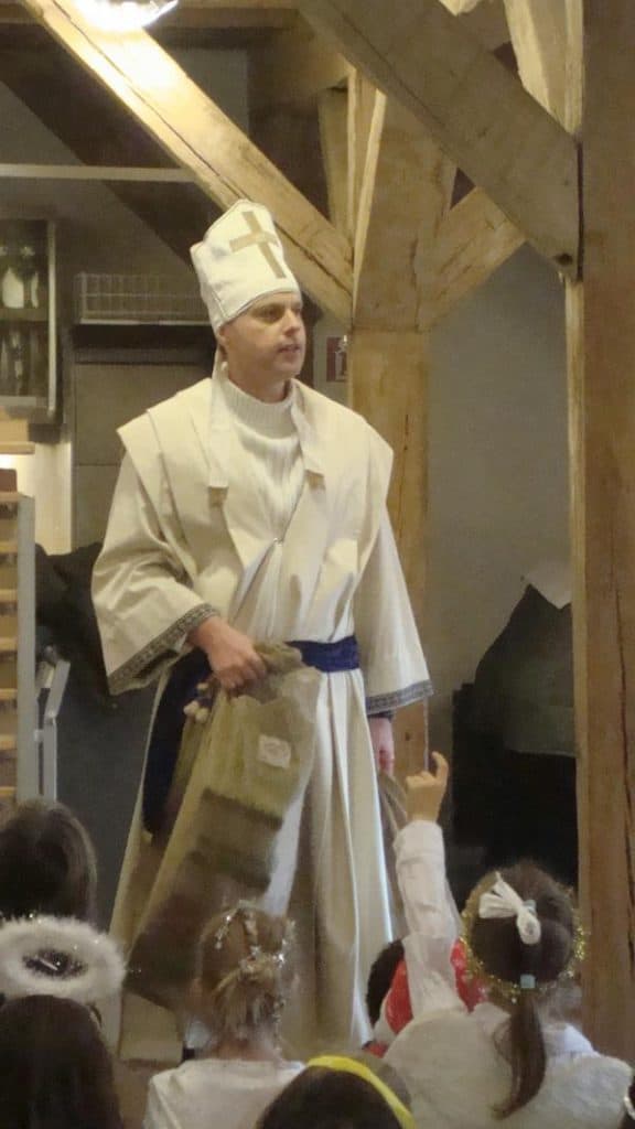 Eine Person in weißem Zeremoniengewand und mit hohem Hut steht im Inneren und hält einen Sack in der Hand. Mehrere Kinder, einige in Engelskostümen und mit Stirnbändern, sitzen davor, beobachten die Person und zeigen auf sie.