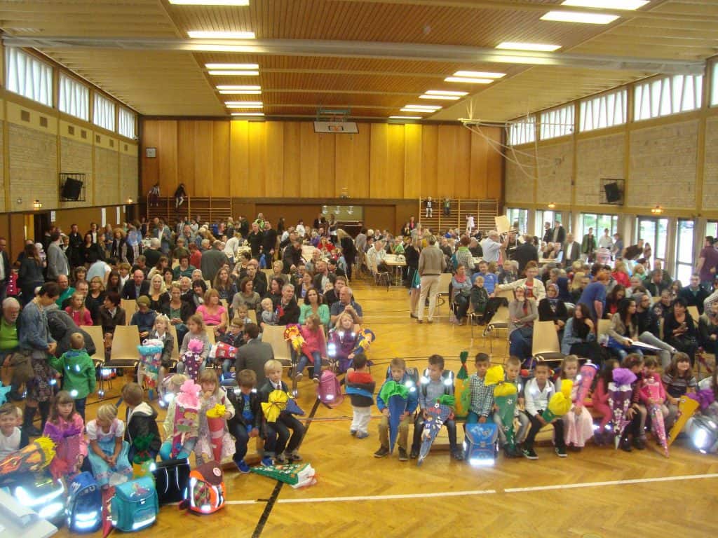 Eine große Gruppe Kinder in bunten Kostümen sitzt in der ersten Reihe einer überfüllten Turnhalle, dahinter sitzen viele Erwachsene an Tischen. Der Raum ist hell, mit Holzböden und hohen Fenstern.