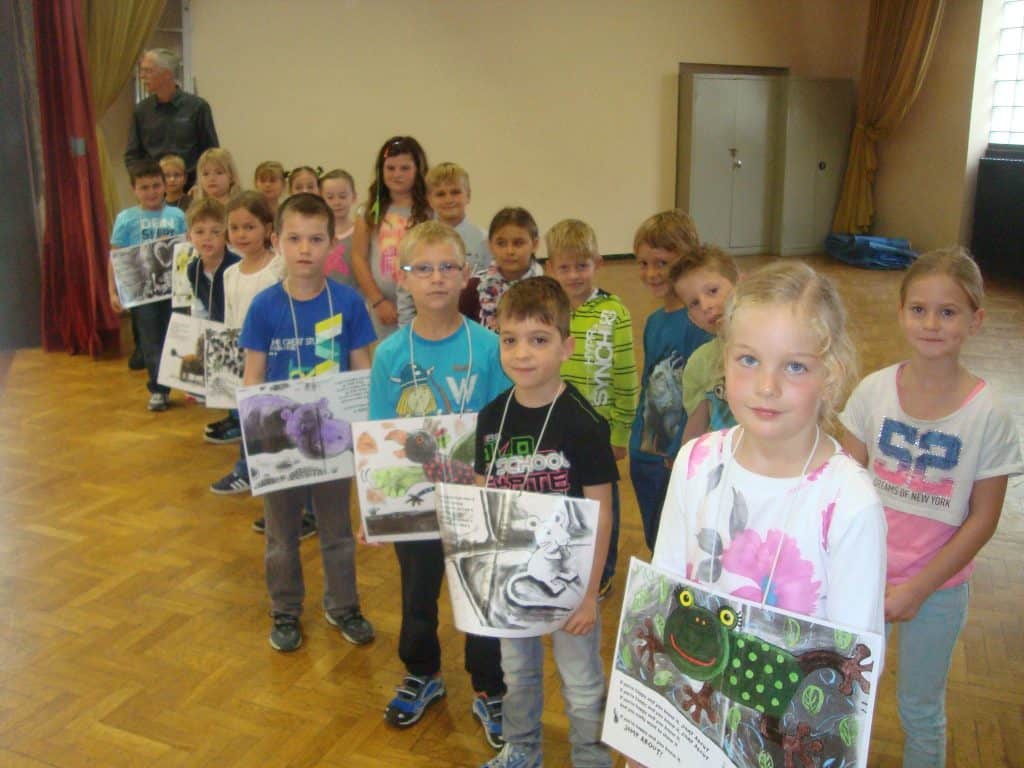 Eine Gruppe von Kindern steht in einer Reihe in einem Raum. Jedes Kind hält ein Poster mit Tier- oder Naturmotiven in der Hand. Die Kinder blicken in die Kamera. Die Szenerie scheint ein Klassenzimmer oder eine Halle mit Holzboden und Vorhängen zu sein.