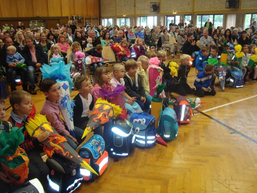 Eine Gruppe kleiner Kinder sitzt auf dem Boden einer Turnhalle und hält jeweils bunte, dekorierte Kegel in der Hand, während ein großes Publikum aus Erwachsenen und Kindern von Sitzen im Hintergrund aus zusieht.