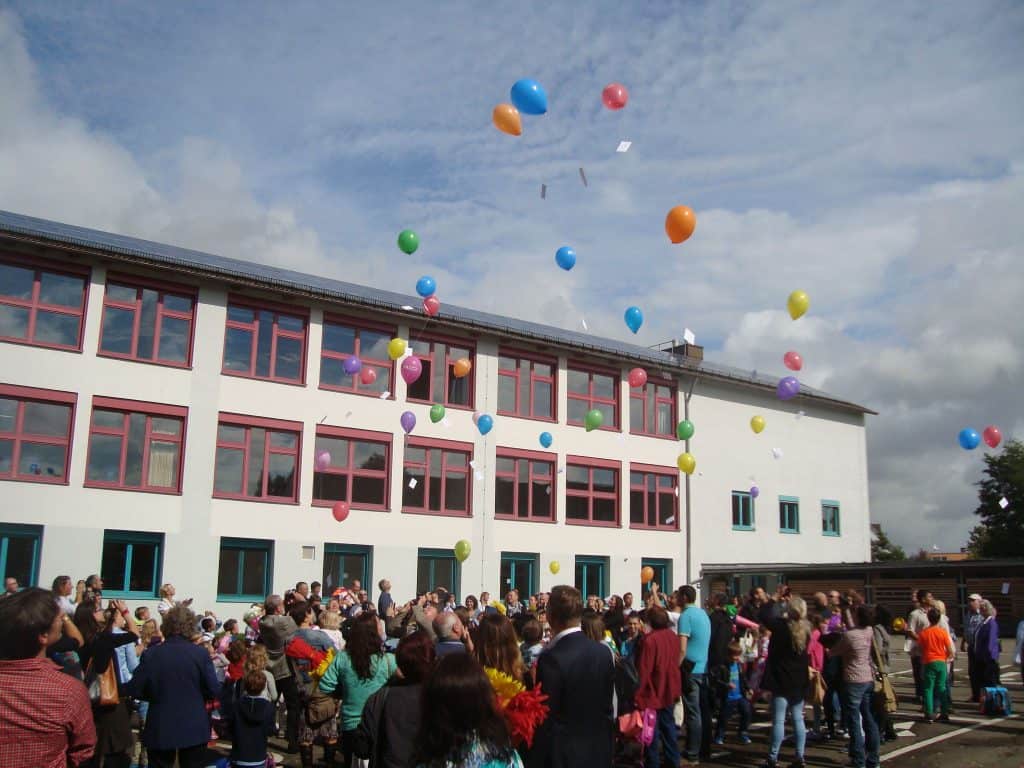 Eine Menschenmenge, darunter Kinder und Erwachsene, versammelt sich vor einem Schulgebäude, während an einem teilweise bewölkten Tag bunte Ballons in den Himmel steigen.