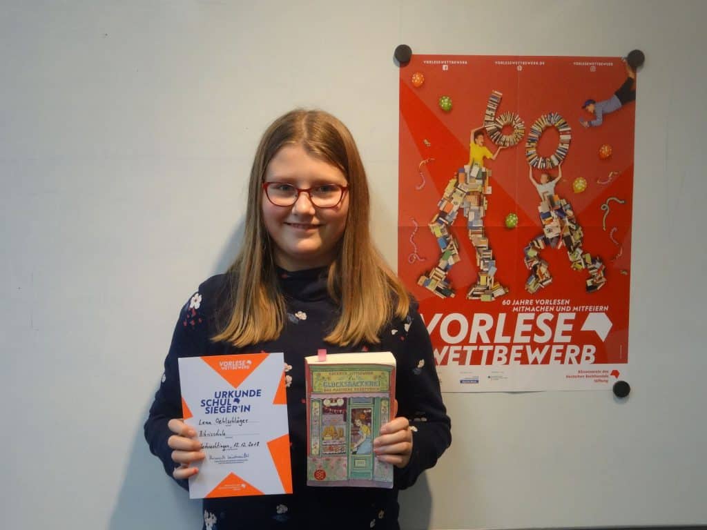 Ein Mädchen mit langen blonden Haaren und roter Brille steht lächelnd da und hält eine Urkunde und ein Buch in den Händen. Hinter ihr an der Wand hängt ein buntes Plakat mit einem deutschen Text über einen Lesewettbewerb.