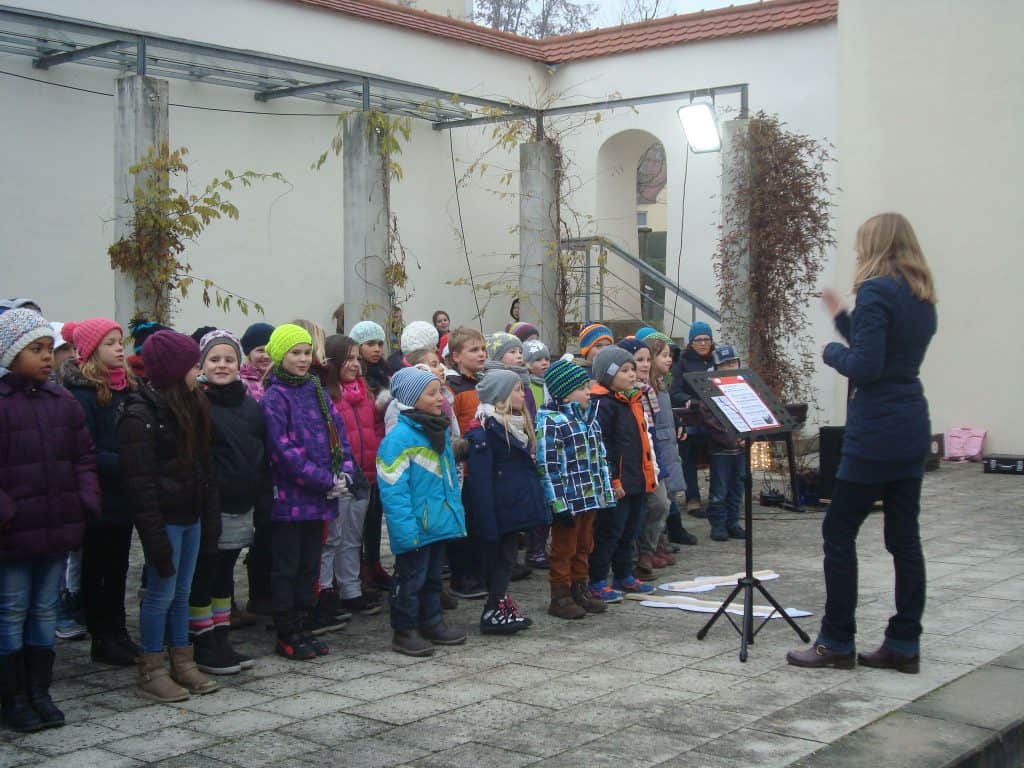 Eine Gruppe Kinder in bunter Winterkleidung singt im Freien, während eine Dirigentin sie dirigiert. Sie stehen auf einer gepflasterten Fläche mit einem Notenständer, umgeben von Säulen und Kletterpflanzen.