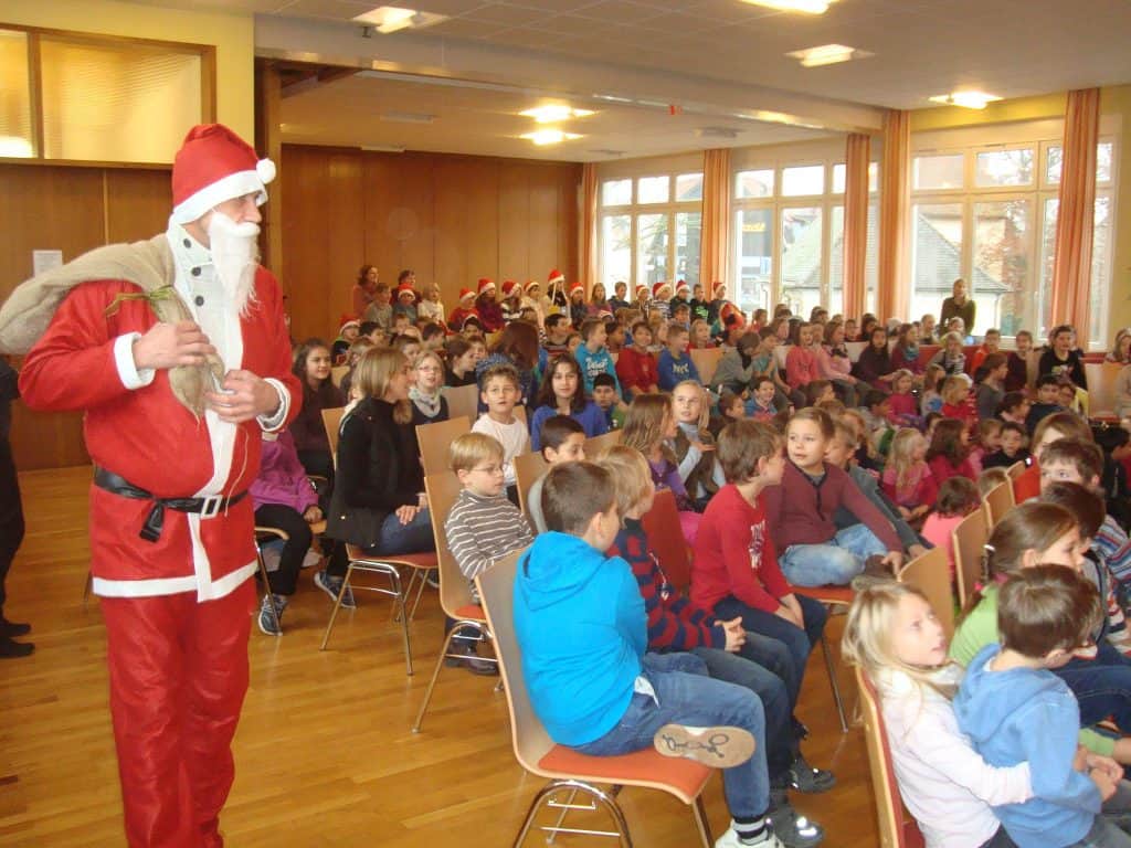 Eine als Weihnachtsmann verkleidete Person steht mit einem Sack in der Hand in einem Raum voller Kinder, die auf Stühlen sitzen und aufmerksam zuschauen. Der Raum ist hell, hat große Fenster und ist für einen festlichen Anlass dekoriert.