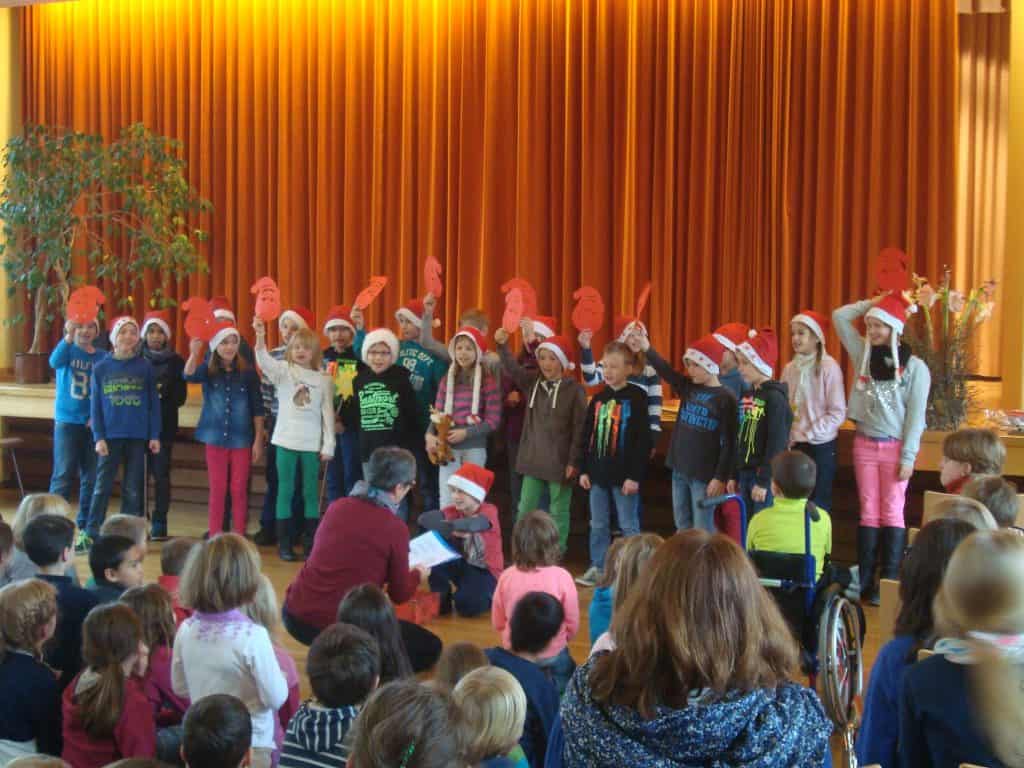 Eine Gruppe von Kindern mit Weihnachtsmannmützen singt auf der Bühne vor einem orangefarbenen Vorhang, während ein Publikum aus Kindern und Erwachsenen zusieht. Einige Kinder auf der Bühne halten rote Papierrequisiten hoch.