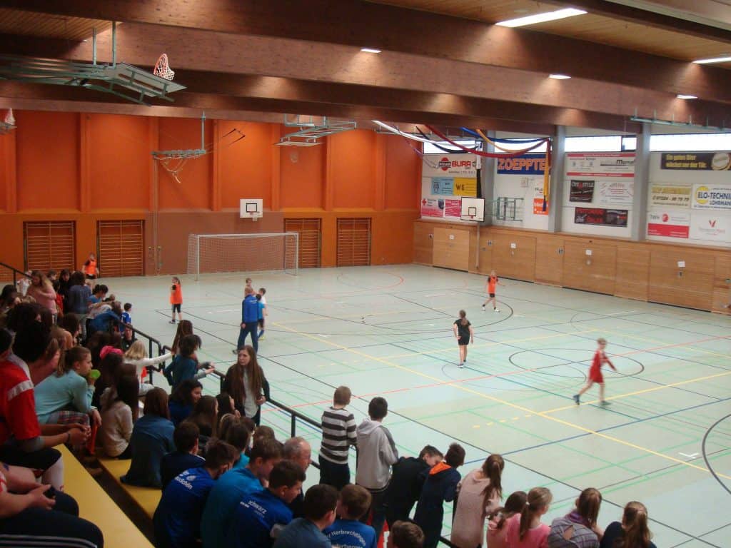 Eine Gruppe Kinder spielt in einer großen Turnhalle ein Hallensportspiel, während Zuschauer von den Tribünen links zuschauen. Die Turnhalle hat hohe Decken, orangefarbene Wände und Basketballkörbe.