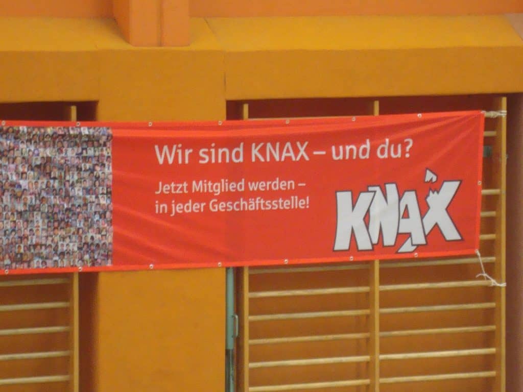 Ein rotes Banner mit deutschem Text: Wir sind KNAX – und du? Jetzt Mitglied werden – in jeder Geschäftsstelle! und ein großes KNAX-Logo, montiert auf einer orangefarbenen Wand, mit kleinen Fotos, die auf der linken Seite eine Collage bilden.