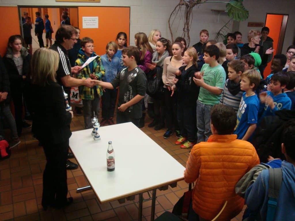 Eine große Gruppe Kinder versammelt sich drinnen um einen Tisch, während ein Junge von einem Erwachsenen einen Händedruck und eine Trophäe erhält, während die anderen zuschauen, klatschen und auf dem gefliesten Boden stehen.