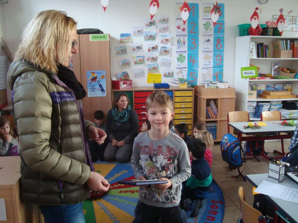 Ein kleiner Junge steht lächelnd mit einem Buch in der Hand in einem Klassenzimmer. Neben ihm steht eine Frau. Andere Kinder und Erwachsene sitzen und arbeiten im Raum, der mit Lehrplakaten und Regalen voller Materialien dekoriert ist.