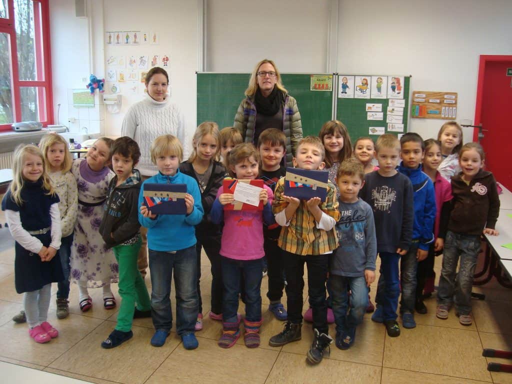 Eine Gruppe kleiner Kinder und zwei Erwachsene stehen zusammen in einem Klassenzimmer. Einige Kinder halten bunte Ordner und Papiere in den Händen. Im Hintergrund sind eine Tafel, Zeichnungen und informative Poster zu sehen.