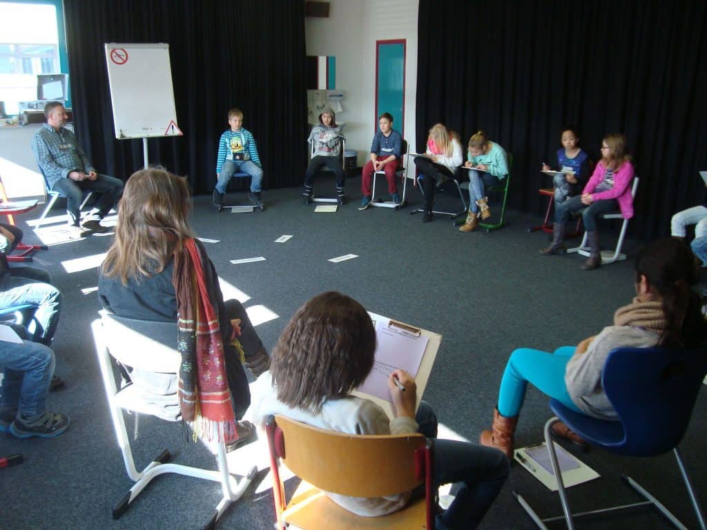 Eine Gruppe von Kindern und ein Erwachsener sitzen in einem Klassenraum im Kreis auf Stühlen. Einige schreiben auf Papier oder Klemmbrettern. Ein Flipchart und verstreute Papiere sind zu sehen und Sonnenlicht fällt durch die Fenster.