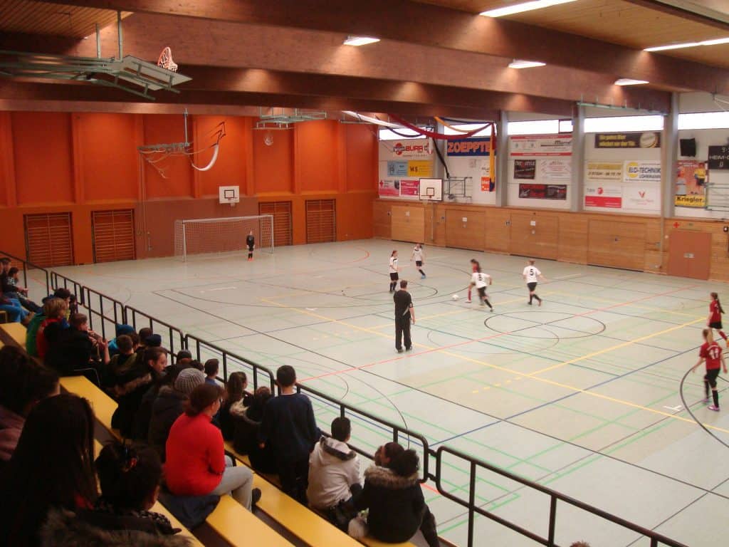 Zuschauer verfolgen ein Hallenfußballspiel in einer Sporthalle. Spieler in rot-weißen Trikots treten auf einem mit Holzwänden und Werbetafeln markierten Spielfeld gegeneinander an. Ein Trainer steht in der Nähe des Spielfelds, darüber sind Basketballkörbe zu sehen.