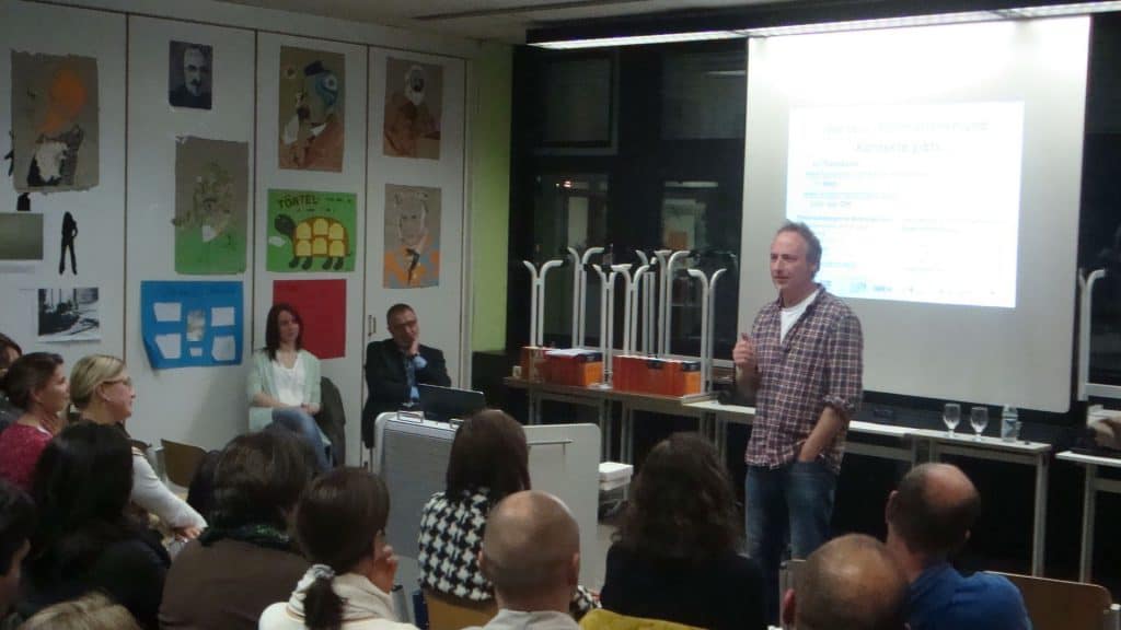 Ein Mann steht und spricht zu einem sitzenden Publikum in einem Klassenzimmer oder Konferenzraum. Hinter ihm läuft auf einer Projektionsleinwand eine Präsentation, und an der Wand hängen bunte Plakate.