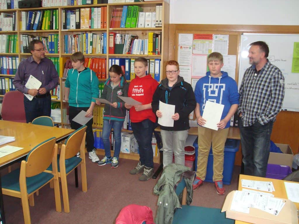 Sechs Schüler stehen mit Papieren in einem Klassenzimmer, flankiert von zwei Erwachsenen. Sie stehen vor Regalen voller Bücher und Ordner. Im Vordergrund stehen Tische und Stühle, an der Wand ist eine Tafel zu sehen.