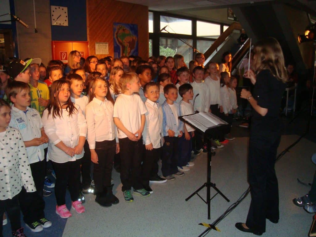 Eine große Gruppe von Kindern, meist in weißen Hemden, steht in einer Schule zusammen und singt, während eine Frau sie dirigiert. Im Raum sind Notenständer und Lichter zu sehen.
