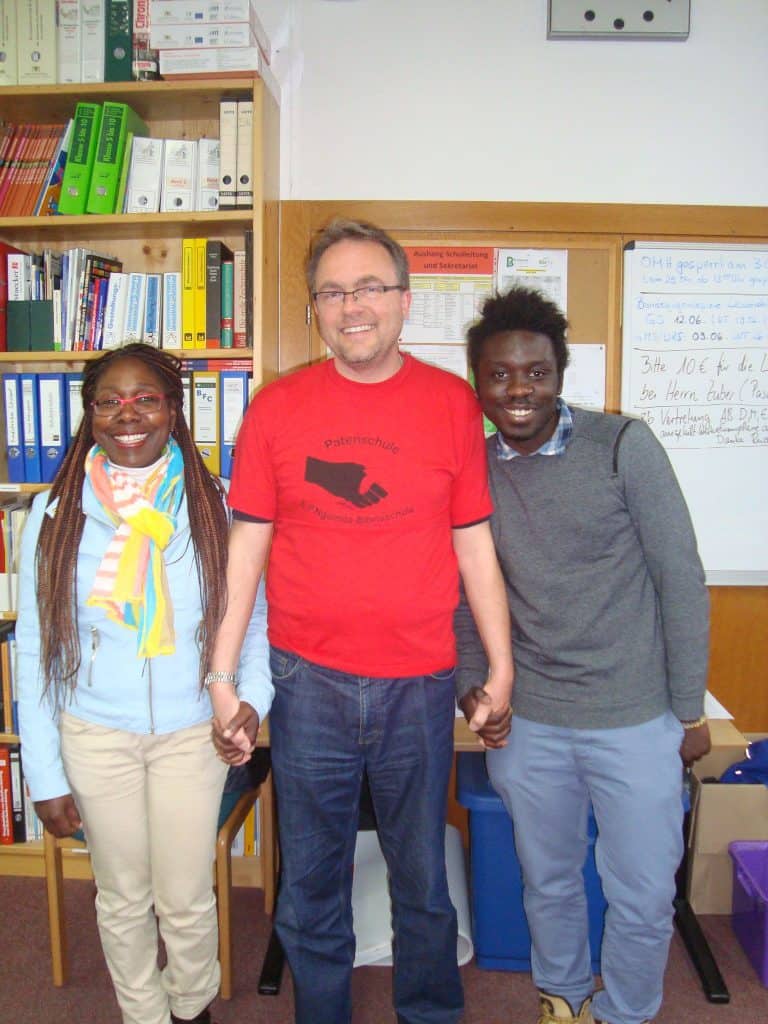 Drei Personen stehen lächelnd im Raum und halten sich an den Händen. Der Mann in der Mitte trägt eine Brille und ein rotes Hemd, die beiden Personen auf beiden Seiten tragen Pullover und Schals. Im Hintergrund sind Bücherregale und eine Tafel zu sehen.