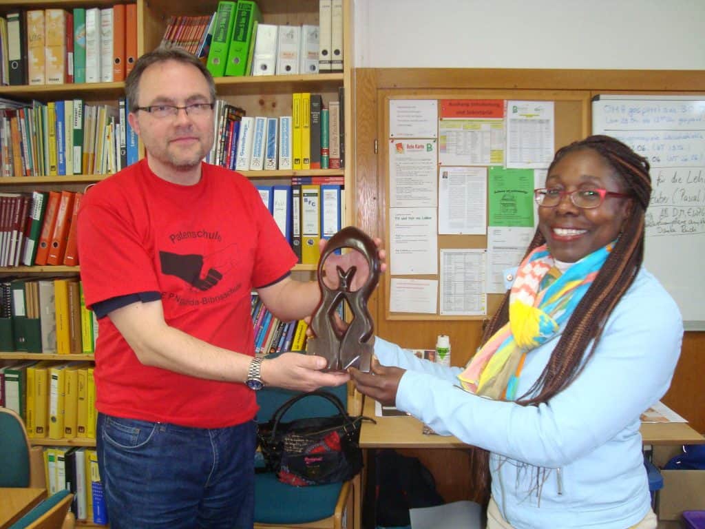 Zwei Personen stehen lächelnd in einem Büro und halten eine geschnitzte Holzauszeichnung in den Händen. Im Hintergrund sind Regale mit Büchern und bunten Ordnern zu sehen.
