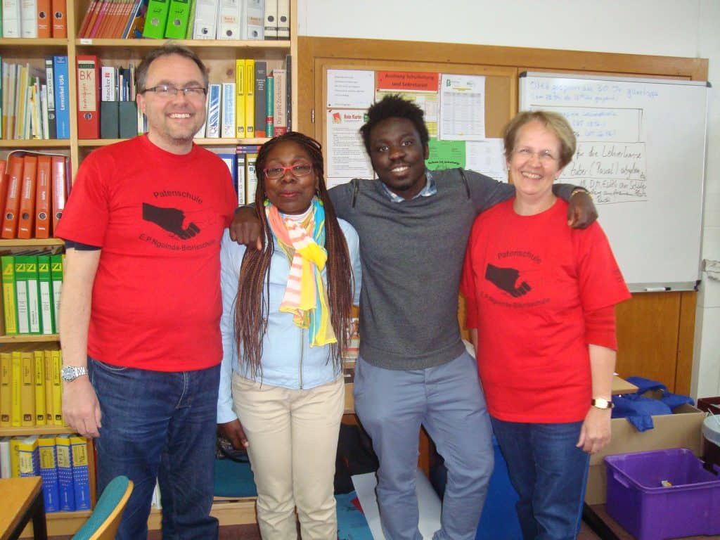 Vier Erwachsene stehen dicht beieinander und lächeln in die Kamera in einem Büro oder Klassenzimmer. Zwei tragen passende rote Hemden, die anderen beiden Freizeitkleidung. Im Hintergrund sind bunte Ordner und eine weiße Tafel zu sehen.
