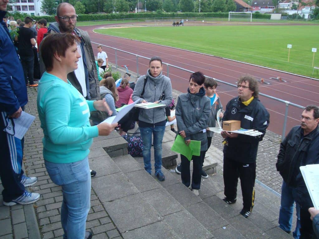 Eine Frau steht mit Papieren auf einer Treppe und spricht zu einer Gruppe Erwachsener, die aufmerksam in der Nähe einer Laufbahn auf einer Sportanlage zuhören. Einige halten Ordner und Kartons in der Hand, andere sitzen in der Nähe.