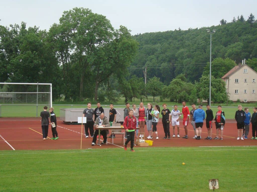 Eine Gruppe von Menschen, hauptsächlich Teenager, steht auf einer Sportbahn neben einem Tisch mit Geräten. Einige stehen in einer Reihe, andere schauen zu oder bereiten sich vor. Im Hintergrund sind grünes Gras, Torpfosten und Bäume zu sehen.