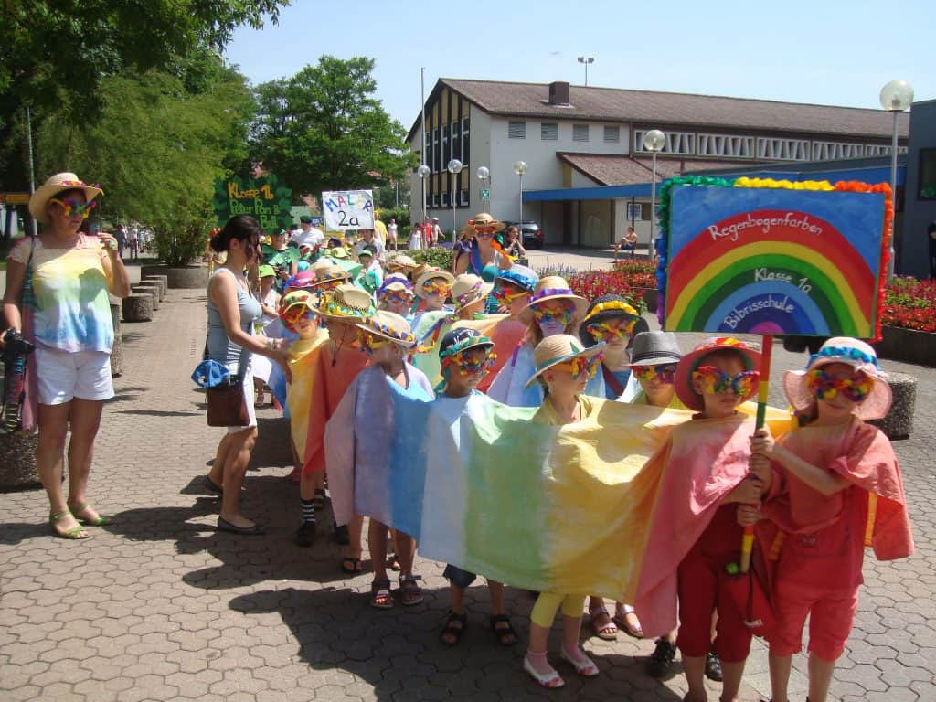 Eine Gruppe von Kindern in regenbogenfarbenen Outfits und Hüten marschiert in einer Parade im Freien. Sie halten Banner mit Regenbögen und Klassenschildern in der Hand und werden von einem Erwachsenen begleitet. Im Hintergrund sind leuchtende Blumen und ein Gebäude zu sehen.