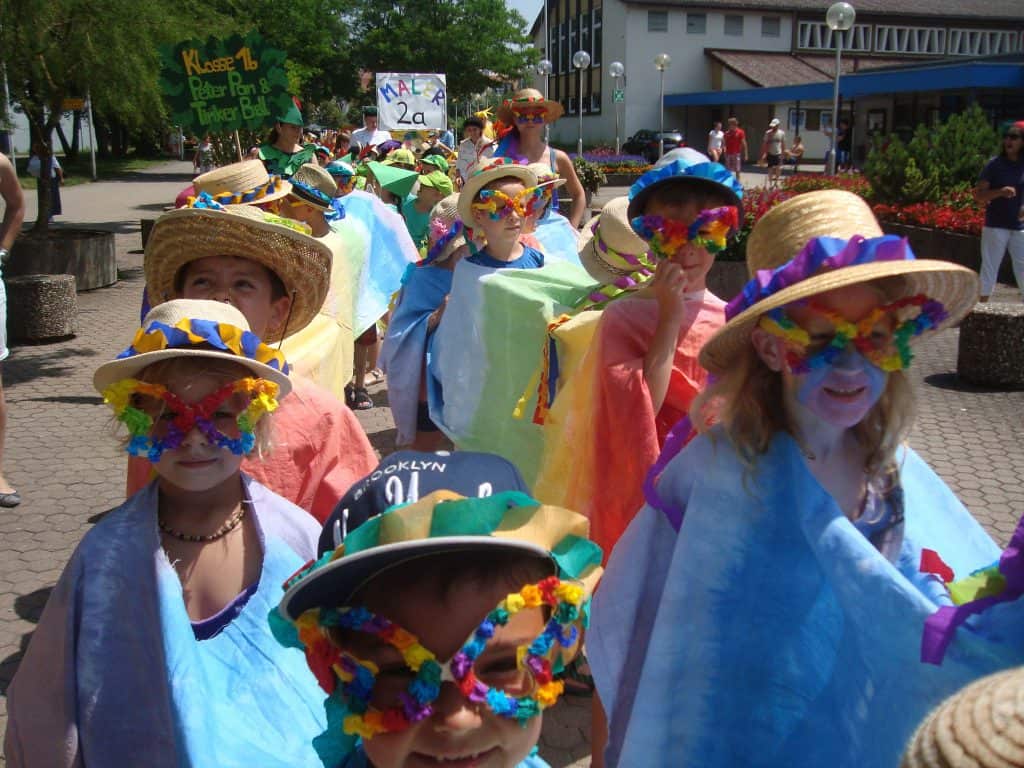 Eine Gruppe Kinder in farbenfrohen Kostümen, Blumengirlanden und Hüten paradiert draußen in der Sonne. Einige halten Schilder, alle lächeln und sind festlich gekleidet, was auf eine Schul- oder Gemeindefeier schließen lässt.