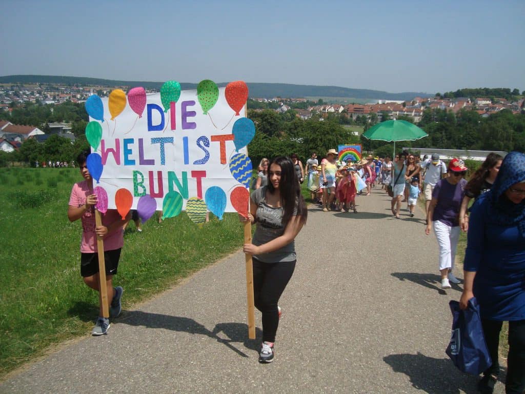 Menschen ziehen in einer Parade durch die Luft. Zwei Personen halten ein buntes Schild mit der Aufschrift „Die Welt ist bunt“. Andere tragen Gegenstände mit Regenbogenmotiven und Regenschirme. Im Hintergrund sind ein Dorf und Hügel zu sehen.