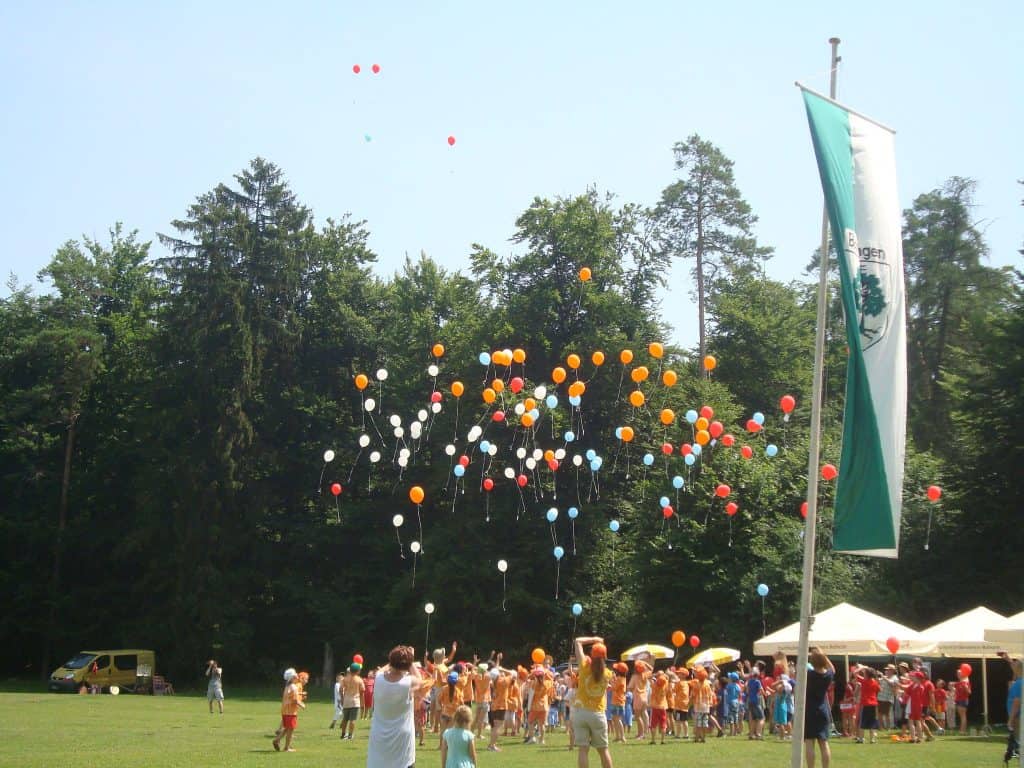 Eine Gruppe von Menschen, darunter auch Kinder, versammelt sich auf einer Wiese und lässt bunte Luftballons in den Himmel steigen. Im Hintergrund sind an einem sonnigen Tag Zelte, eine Flagge und Bäume zu sehen.