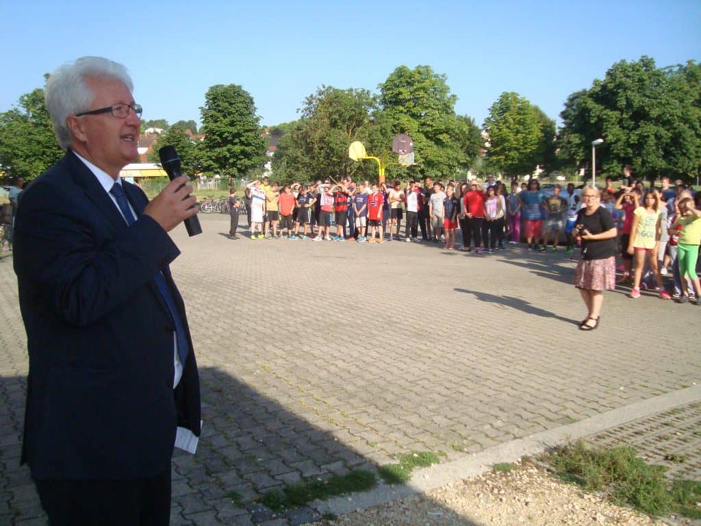 Ein Mann im Anzug spricht im Freien in ein Mikrofon, während im Hintergrund eine Gruppe von Kindern und Erwachsenen auf einer gepflasterten Fläche in der Nähe von Bäumen und einem Basketballkorb steht.