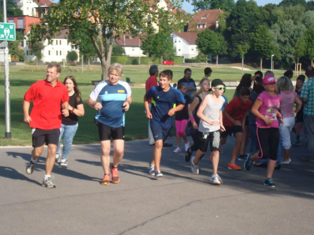 Eine Gruppe von Menschen, darunter Kinder und Erwachsene, läuft an einem sonnigen Tag im Freien. Sie scheinen an einem Spaßlauf oder Rennen teilzunehmen. Im Hintergrund sind grüne Bäume und Gebäude zu sehen.