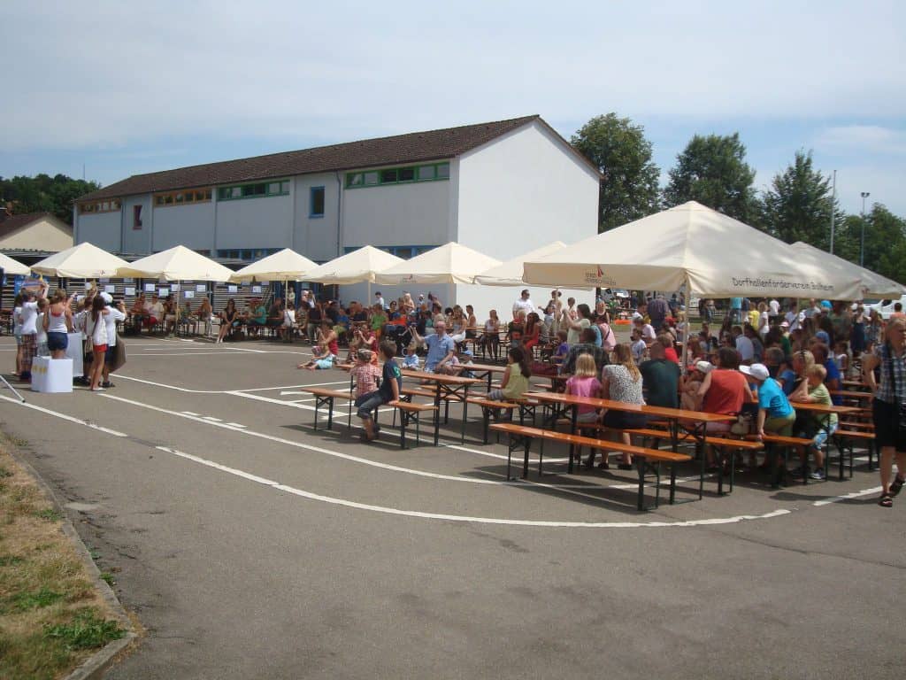 Eine Gemeinschaftsveranstaltung im Freien: Menschen sitzen an langen Holztischen unter großen Zelten auf einem Parkplatz in der Nähe eines weißen Gebäudes. Andere laufen herum und versammeln sich an Ständen. Das Wetter ist sonnig.
