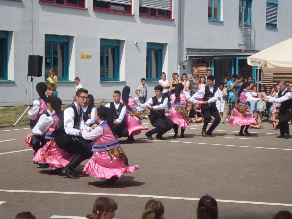 Eine Gruppe junger Leute in traditioneller Kleidung führt im Freien einen Volkstanz auf. Männer heben Frauen in rosa Röcken hoch. Eine Menge Schaulustiger, darunter auch Kinder, beobachtet die Szene in der Nähe eines Gebäudes.