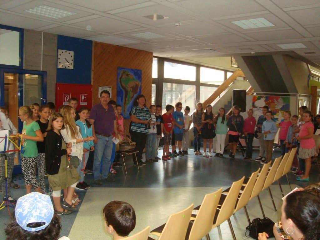 Eine Gruppe von Kindern und einige Erwachsene stehen im Halbkreis in einem hell erleuchteten Raum, andere sitzen ihnen gegenüber in Stuhlreihen. Der Raum hat große Fenster und bunte Poster an den Wänden.