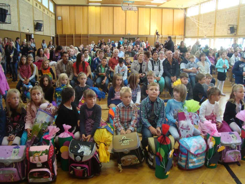 Eine große Gruppe von Kindern sitzt mit dekorierten Schultüten und Rucksäcken in den vorderen Reihen einer Turnhalle, während dahinter viele Erwachsene sitzen, was auf eine Schulveranstaltung oder Feier schließen lässt.
