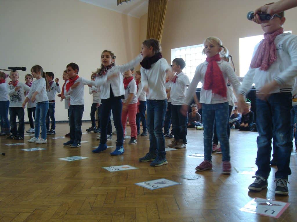 Eine Gruppe von Kindern in Jeans und weißen Hemden, einige mit roten Schals, steht in einer Reihe auf einem Holzboden im Innenbereich und führt eine Aufführung auf. Ein Kind hält ein Fernglas; vor jedem Kind liegen nummerierte Zettel.