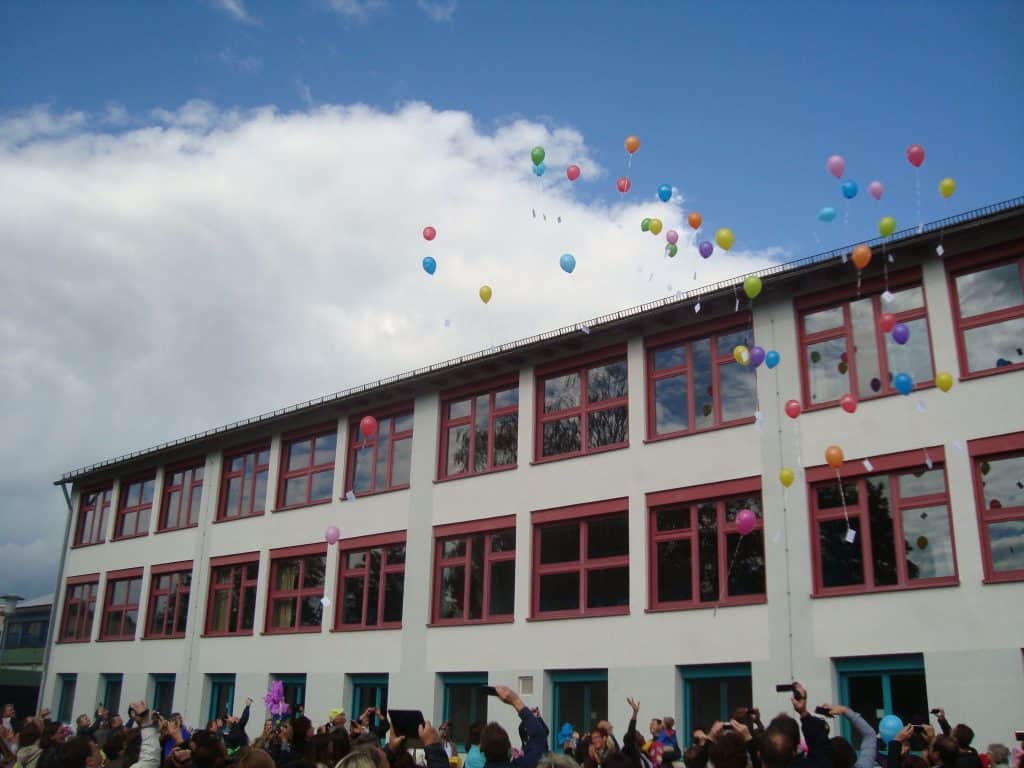 An einem teilweise bewölkten Tag versammelt sich eine Menschenmenge vor einem Schulgebäude, während bunte Ballons in den Himmel steigen.