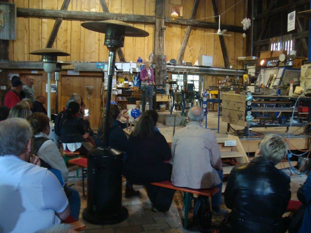 Eine Gruppe von Menschen sitzt auf Bänken in einer rustikalen Holzwerkstatt und beobachtet eine Person, die vorne spricht. Verschiedene Werkzeuge, Maschinen und Heizgeräte sind im Raum verteilt und schaffen eine ungezwungene, kreative Atmosphäre.