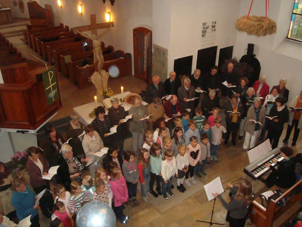 Ein großer Chor aus Erwachsenen und Kindern singt gemeinsam in einer Kirche, begleitet von einem Pianisten. Die Kirche ist mit Kerzen, einem Kreuz und herbstlichen Erntegaben geschmückt. Die Menschen sitzen in den Kirchenbänken und stehen mit Liederbüchern in den Händen.