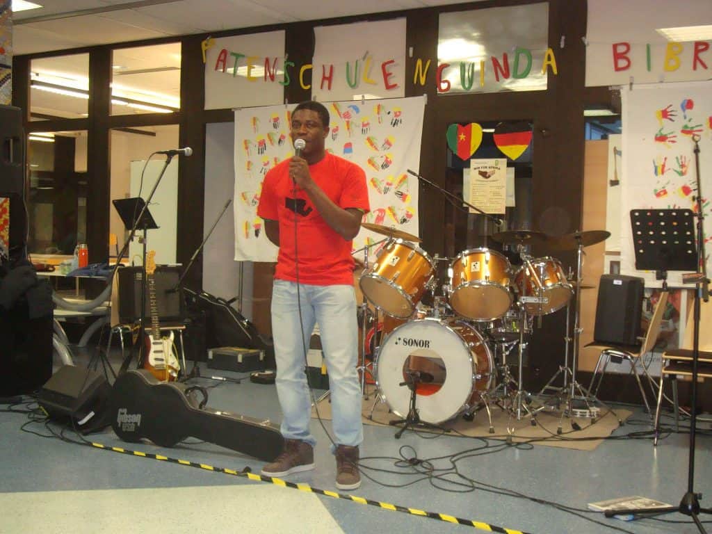 Ein Mann in einem roten Hemd singt in ein Mikrofon auf einer Indoor-Bühne. Hinter ihm stehen Schlagzeug, Gitarren und Notenpulte. Bunte Banner und Plakate schmücken den Hintergrund.