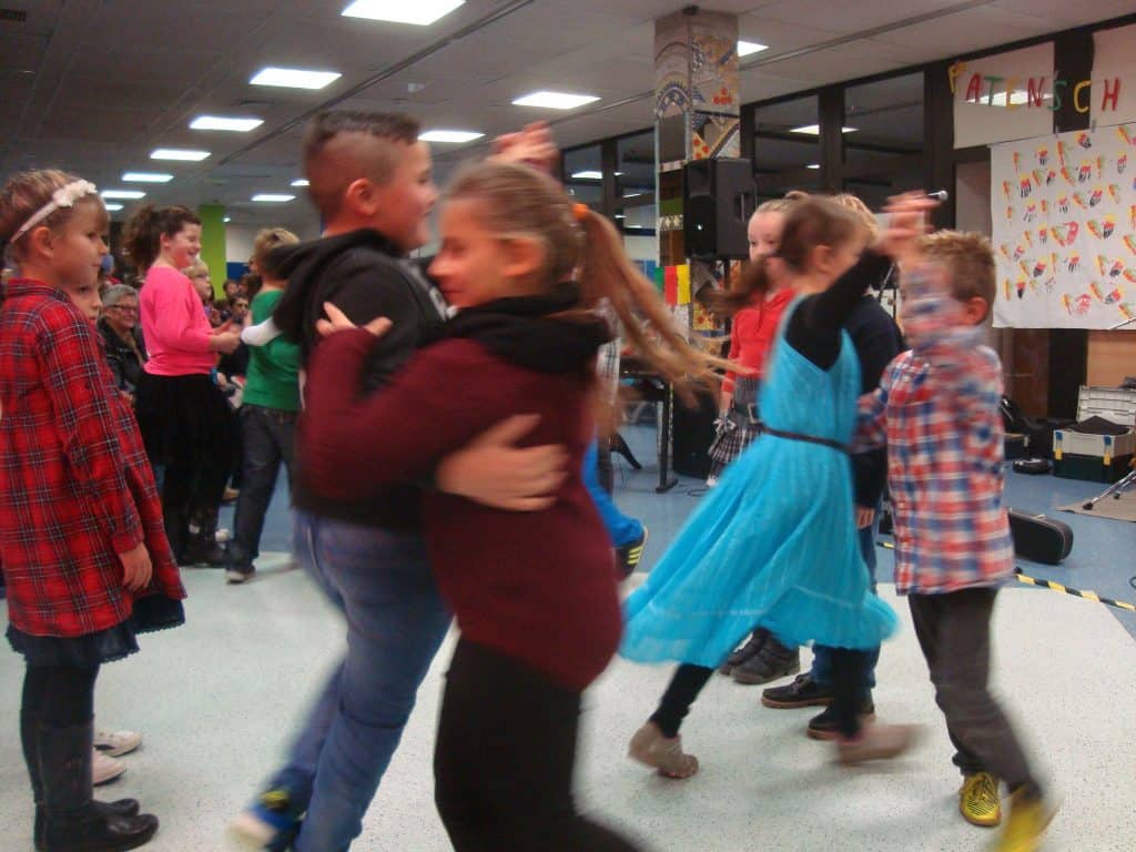 Kinder tanzen paarweise im Haus, manche wirbeln herum. Die Szene ist lebhaft, mit mehreren Kindern in bunter Kleidung. Ein Mädchen in einem blauen Kleid und ein Junge in einem karierten Hemd stehen im Vordergrund. Andere Kinder schauen aus dem Hintergrund zu.